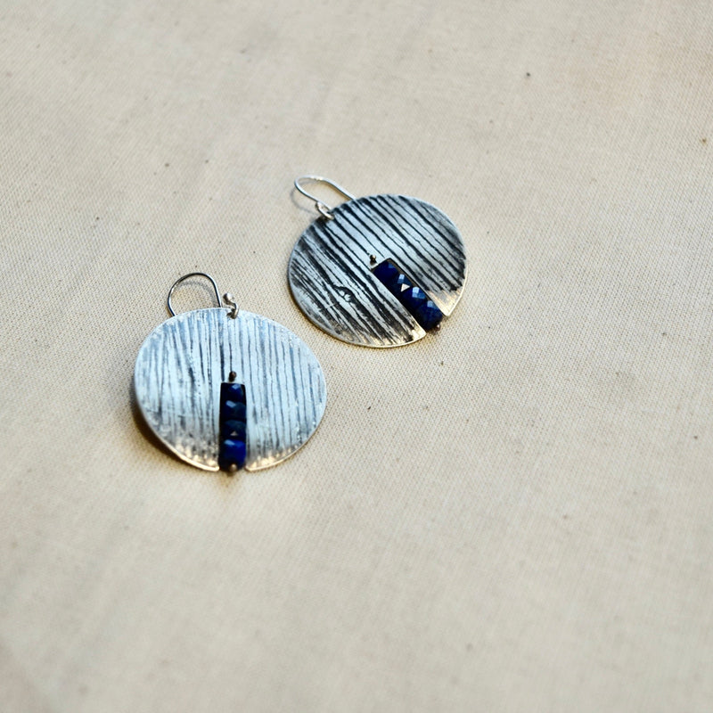 Round earrings with textured design and blue accents on a beige fabric background